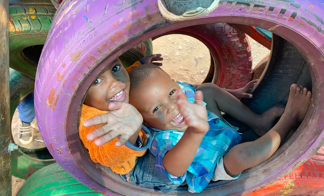 Zwei kleine Jungs spielen in einem buntbemalten Reifen und winken lachend ins Bild. 