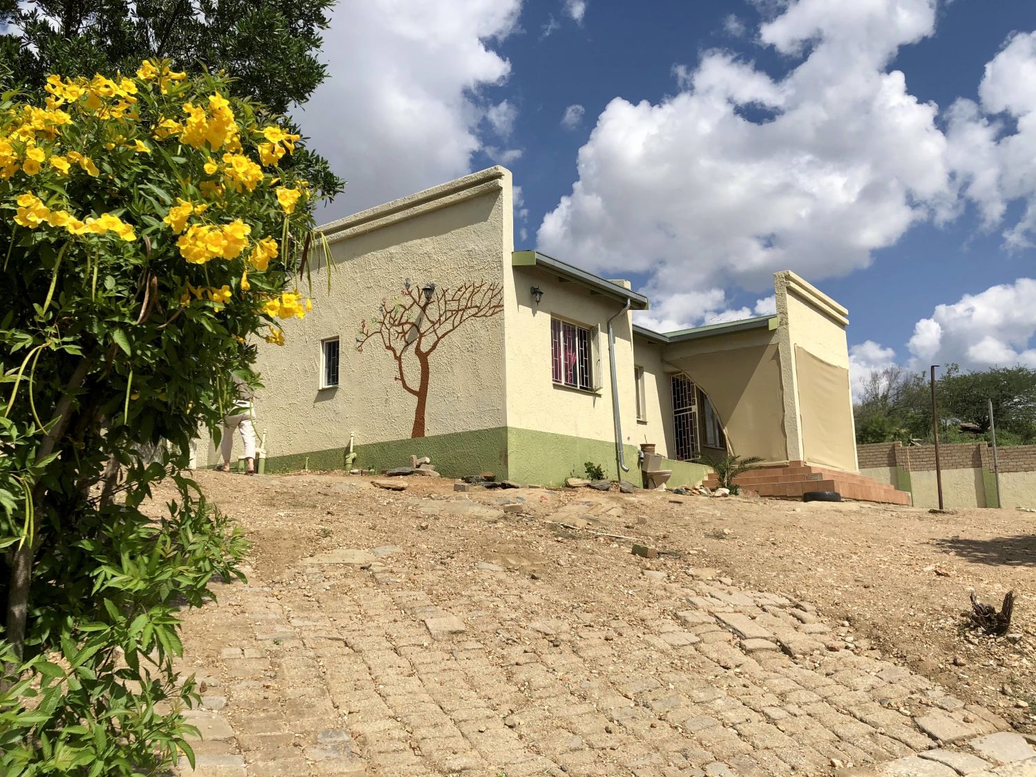Das frisch gestrichene grün-gelbe Haus ist umgeben von eine schützenden Mauer. An der Seite des Hauses ist ein Baum gemalt worden und die großen Büsche im Garten tragen große gelbe Blumen.