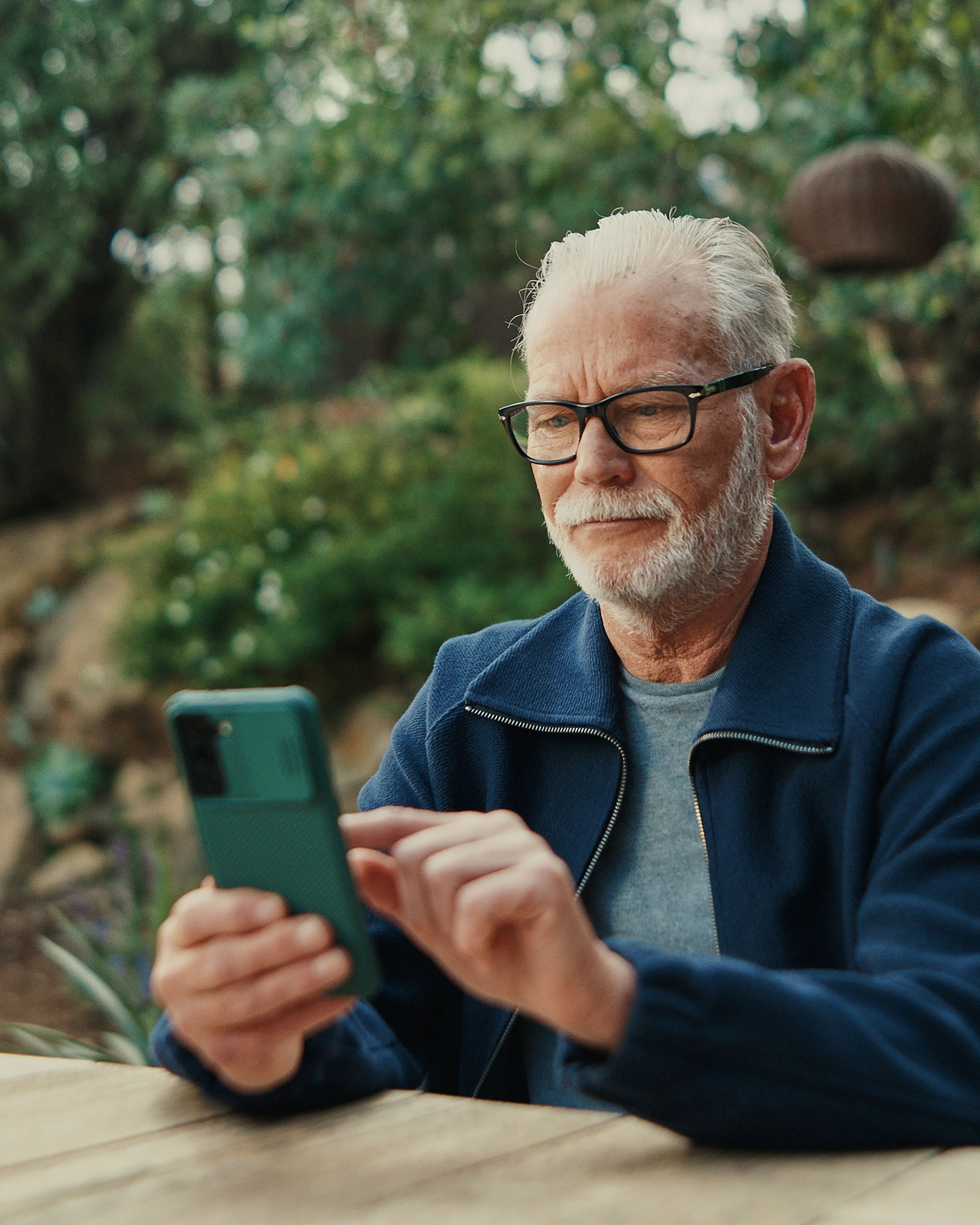 An older man with glasses sitting and using his smart phone.