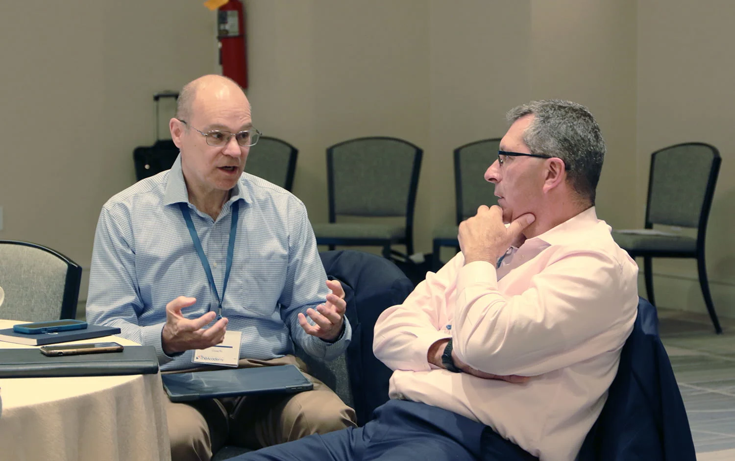 Two men in business attire sitting at a conference table, with one gesturing as he speaks and the other listening attentively with his hand on his chin.