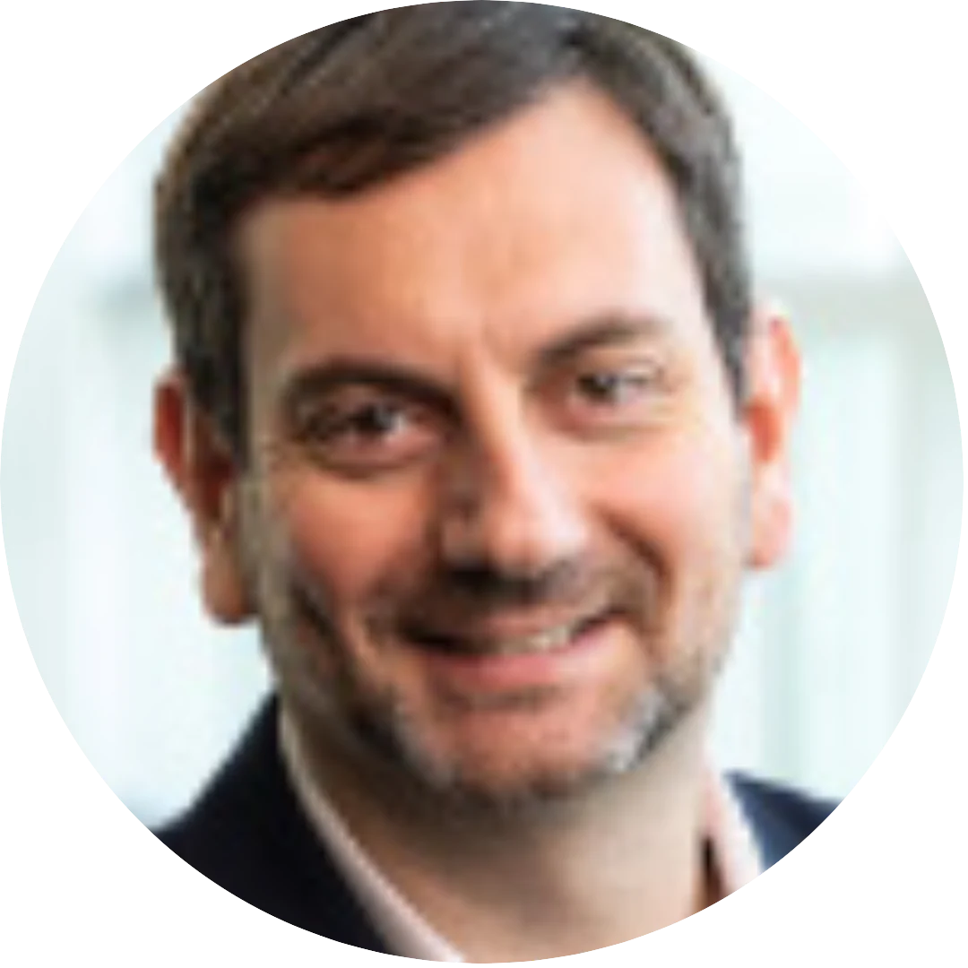 Close-up headshot of a man with short dark hair and facial hair, smiling slightly and wearing a suit jacket and collared shirt, set against a softly blurred indoor background.