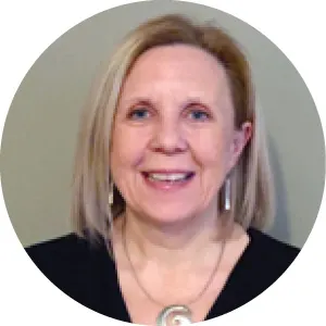 Smiling woman with straight blonde hair wearing a black top and a silver spiral pendant necklace, posed in front of a neutral gray background.