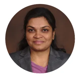 A woman with long dark hair wearing a gray blazer and purple top smiles slightly in a professional headshot against a solid maroon background.