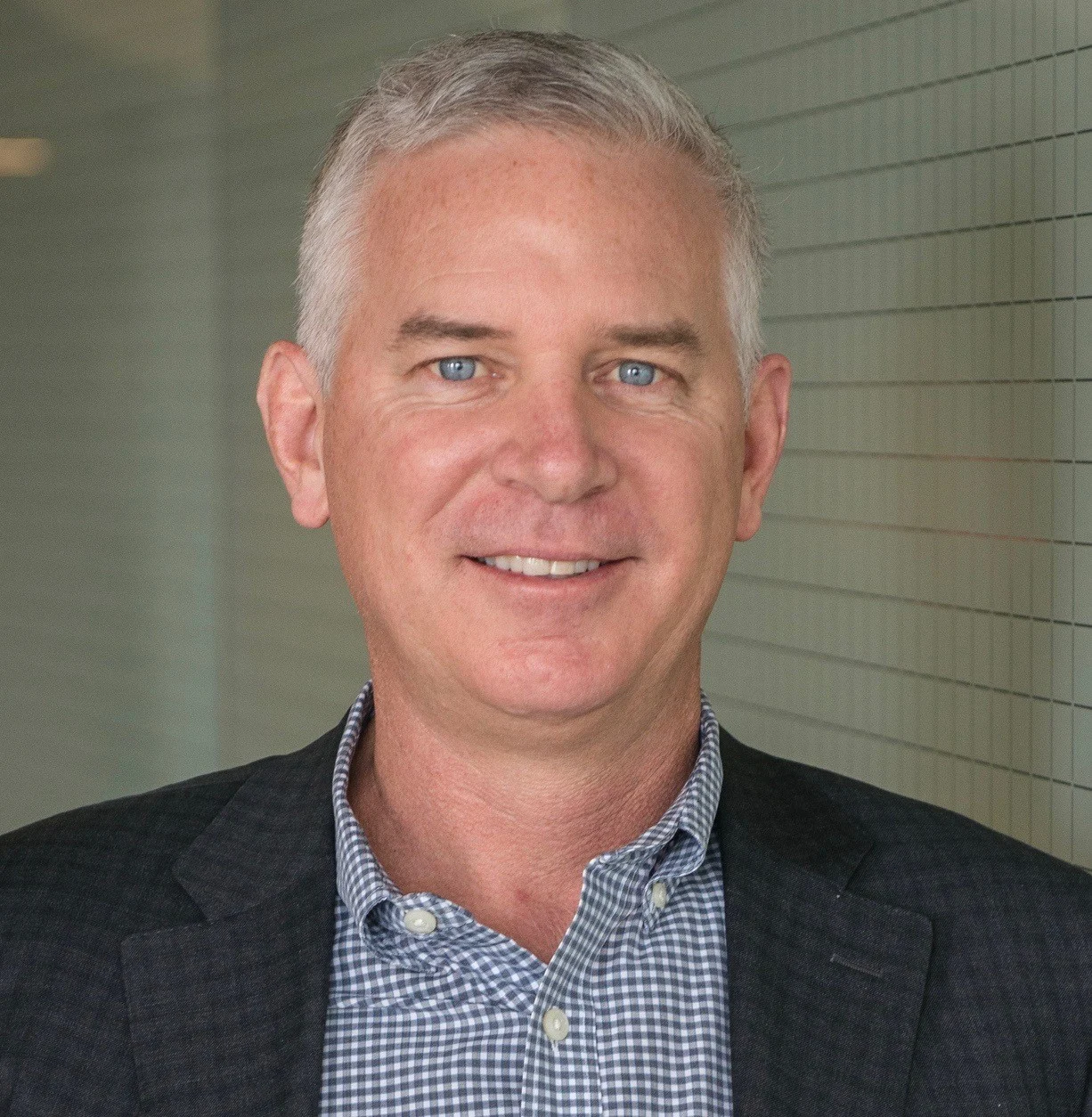 A professional headshot of a man with short gray hair wearing a checkered shirt and dark blazer, standing in front of a tiled wall background.