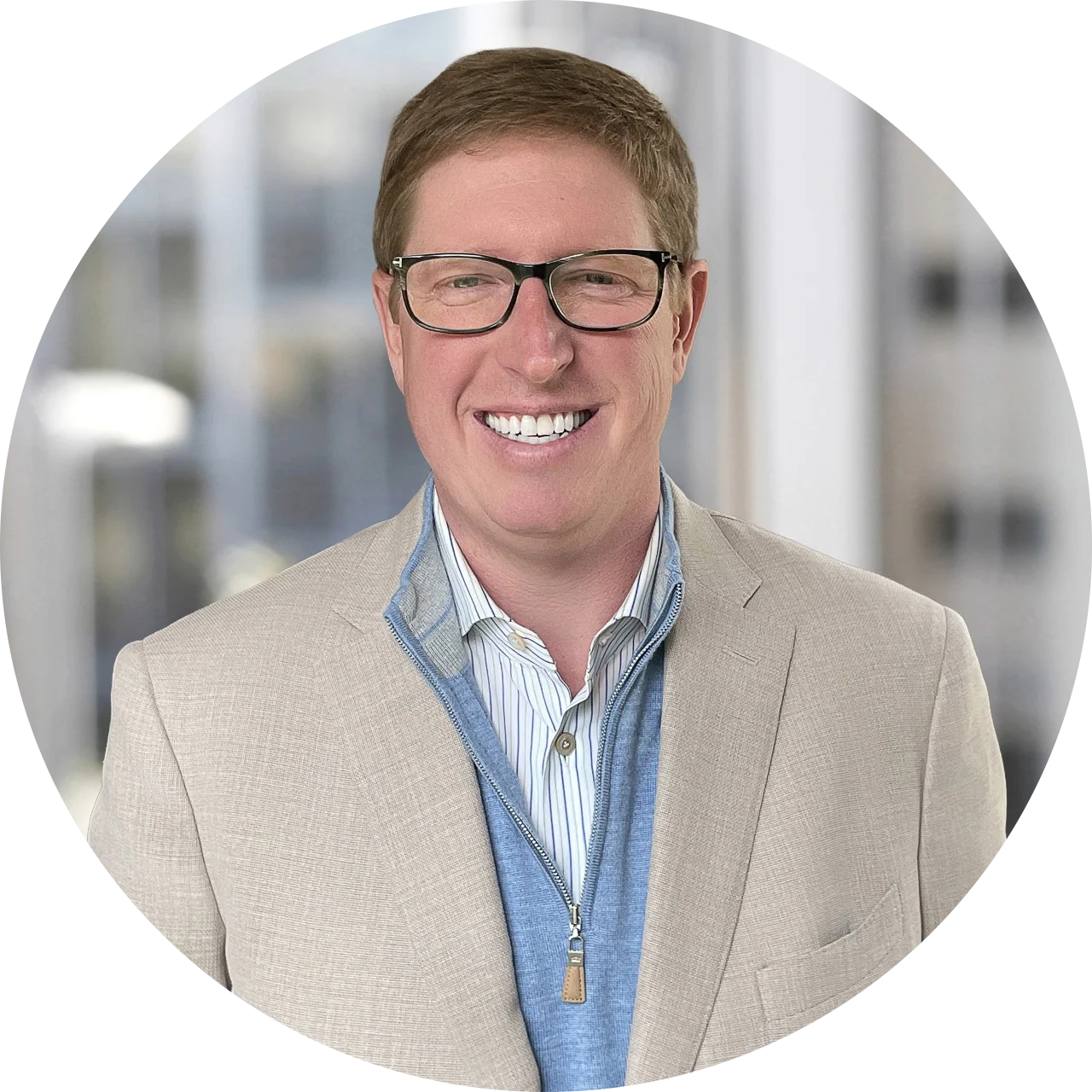 A smiling man wearing glasses, a beige blazer, a blue zip-up sweater, and a striped shirt, standing in front of a blurred office window background.