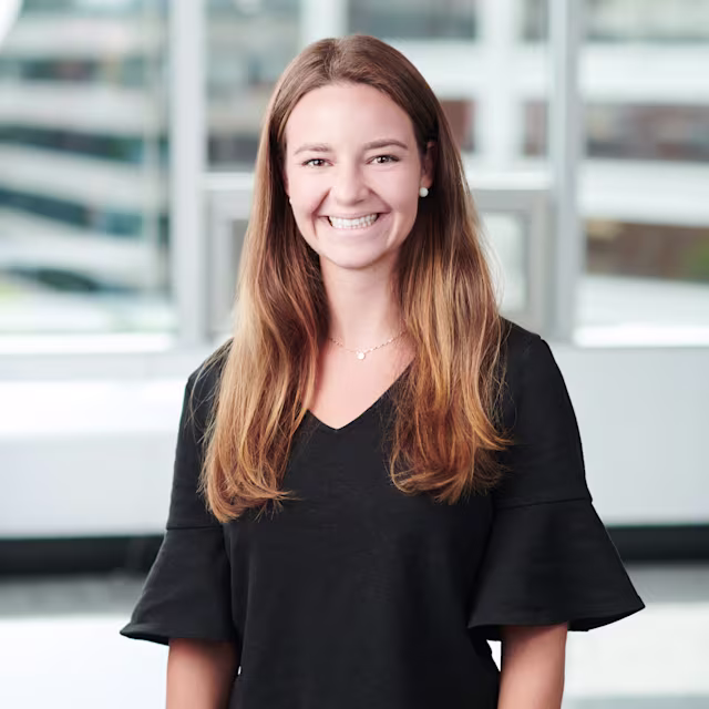 Smiling woman with long light brown hair wearing a black V-neck top with ruffled sleeves, standing in a modern office setting with large windows in the background.
