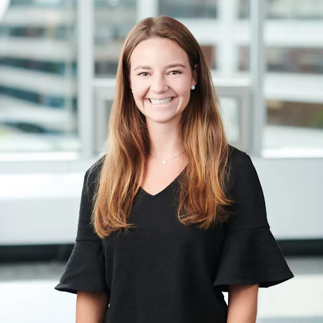Smiling woman with long light brown hair wearing a black V-neck top with ruffled sleeves, standing in a modern office setting with large windows in the background.