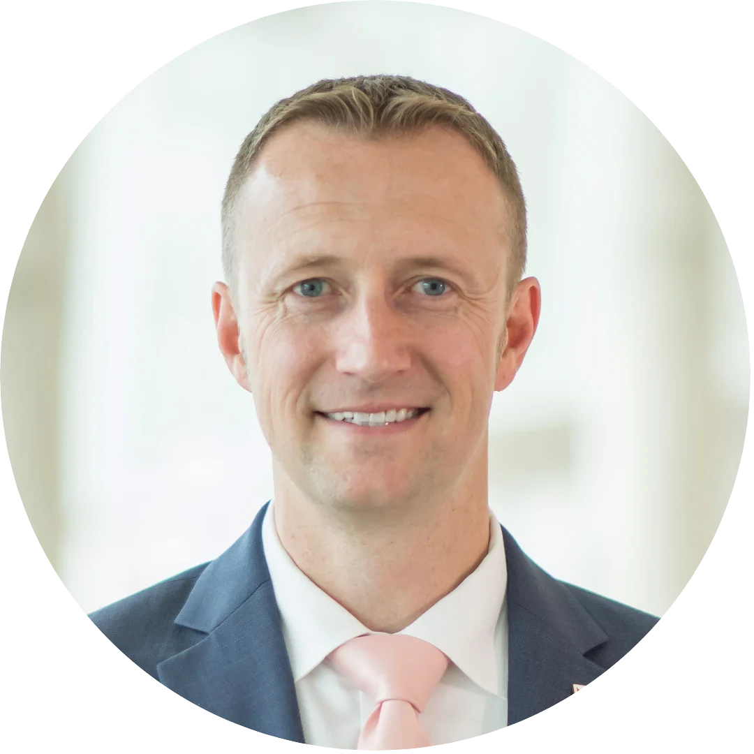 Smiling man wearing a navy suit, white shirt, and light pink tie, posed in front of a softly blurred bright indoor background.