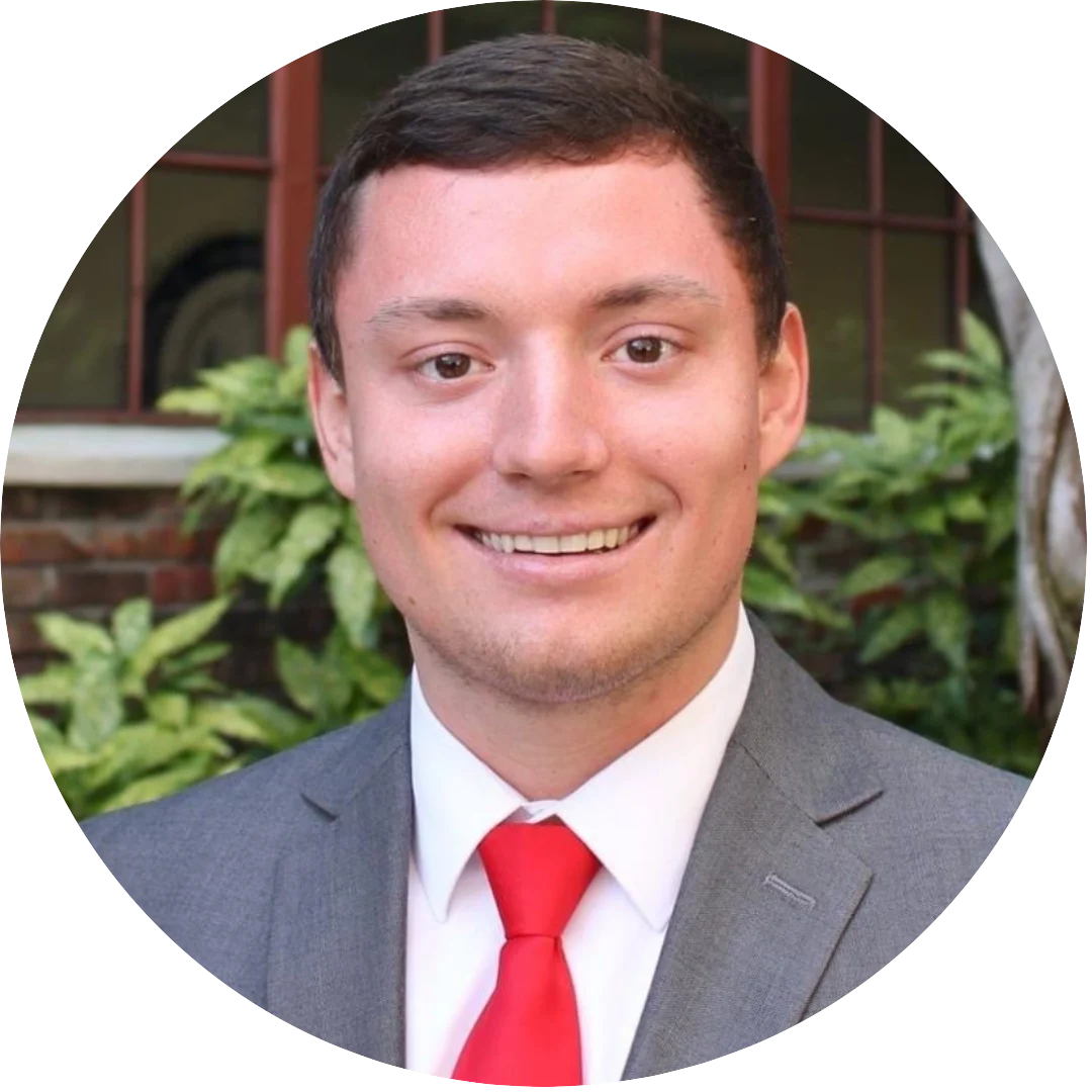 Smiling man wearing a gray suit, white shirt, and bright red tie, posed outdoors in front of greenery and a brick building with windows.