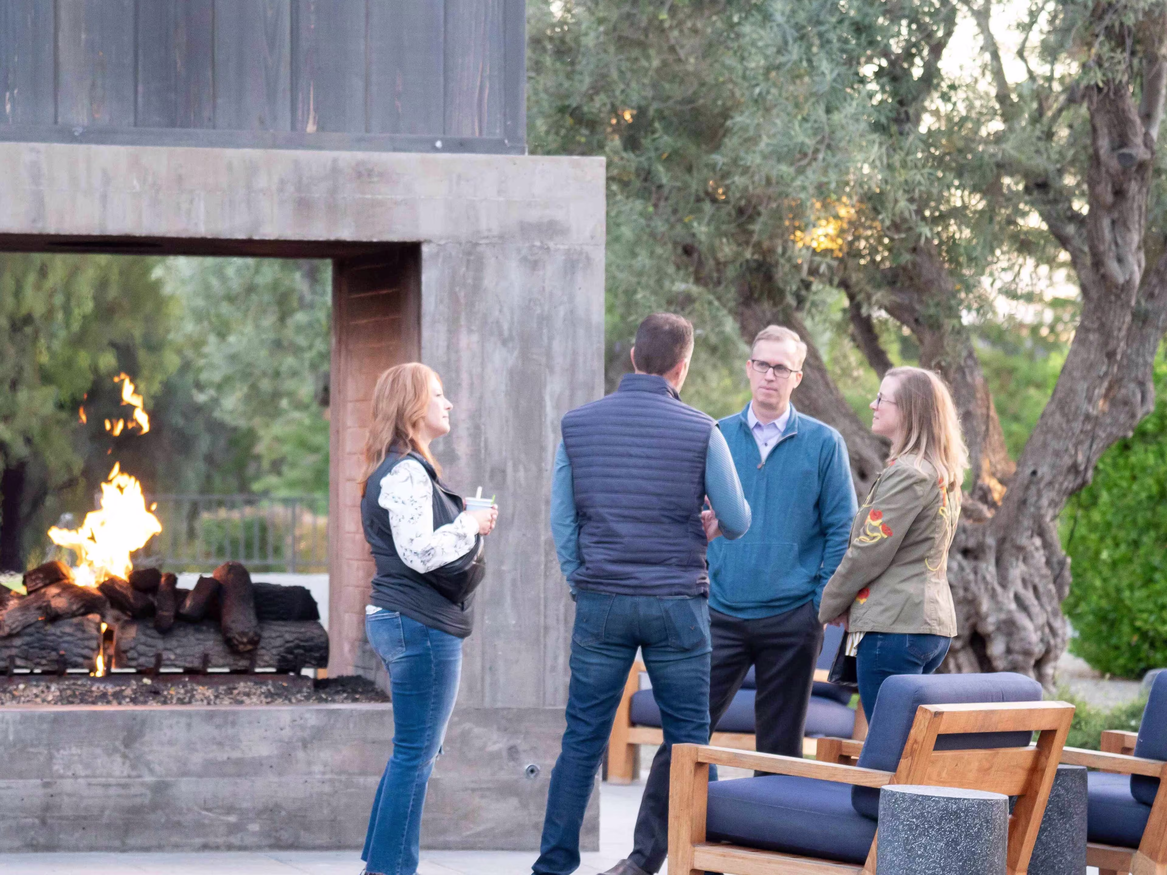A small group of professionals talk outdoors beside a modern open-air fireplace during a healthcare financial management conference, with trees and lounge seating in the background.