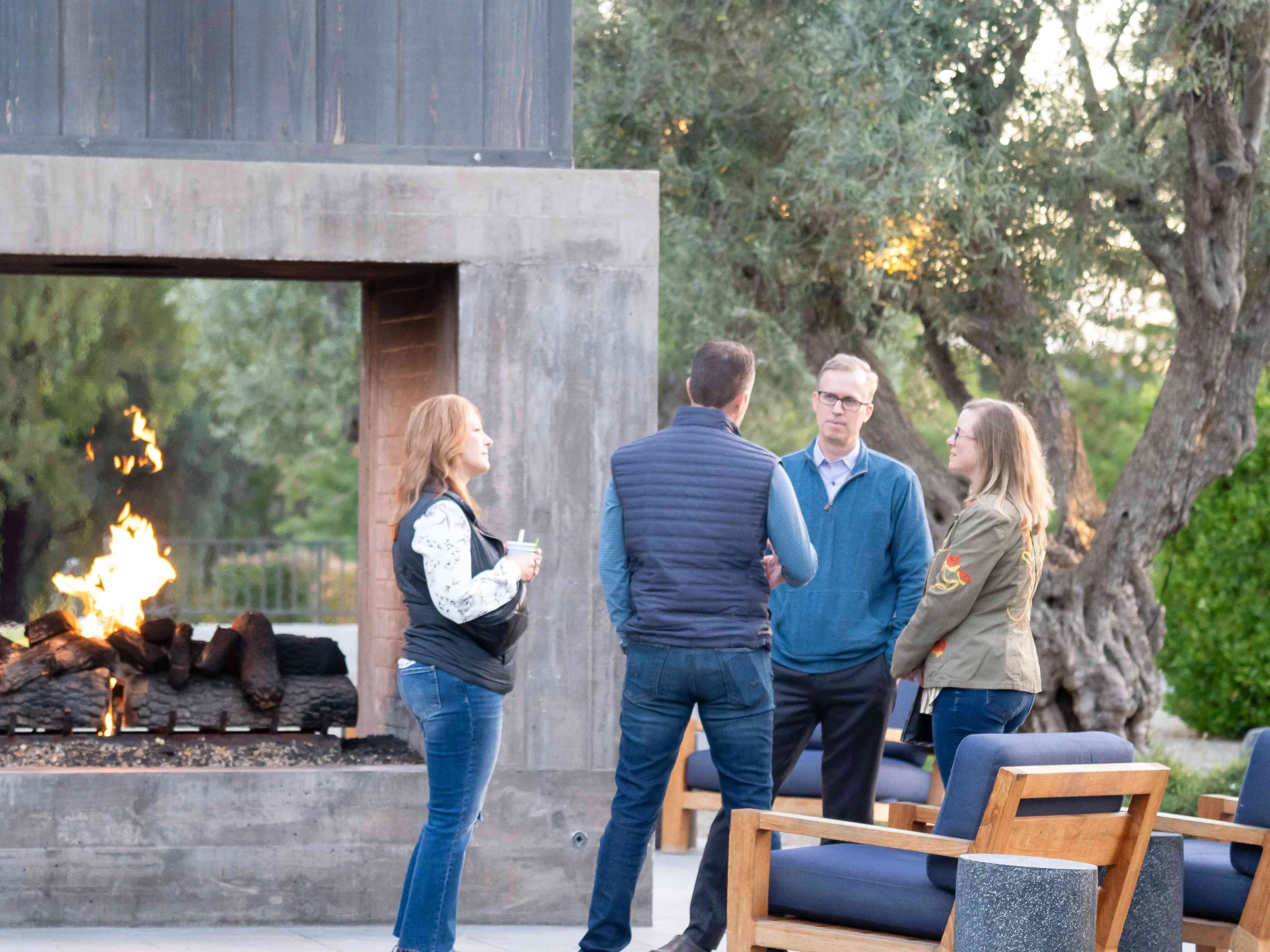 A small group of professionals talk outdoors beside a modern open-air fireplace during a healthcare financial management conference, with trees and lounge seating in the background.
