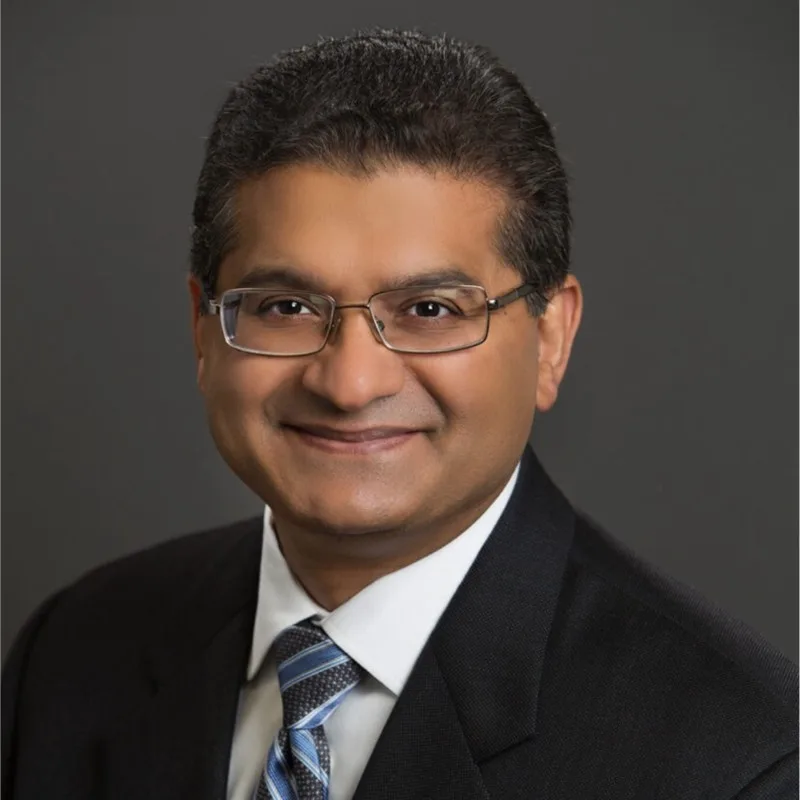 A man wearing a dark suit, striped tie, and glasses smiles at the camera in a formal headshot set against a solid dark background.