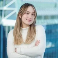 A woman with long, straight blonde hair wearing a white sweater poses confidently with arms crossed in front of a blurred modern building background. 