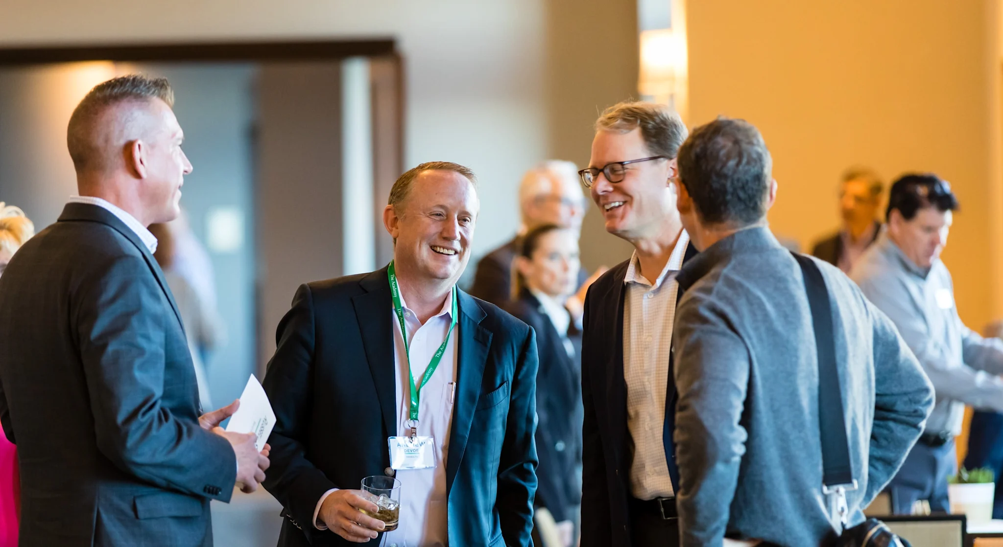 Group of professionally dressed individuals engaged in conversation at a networking event. Participants appear smiling and relaxed in a well-lit venue with others interacting in the background.







