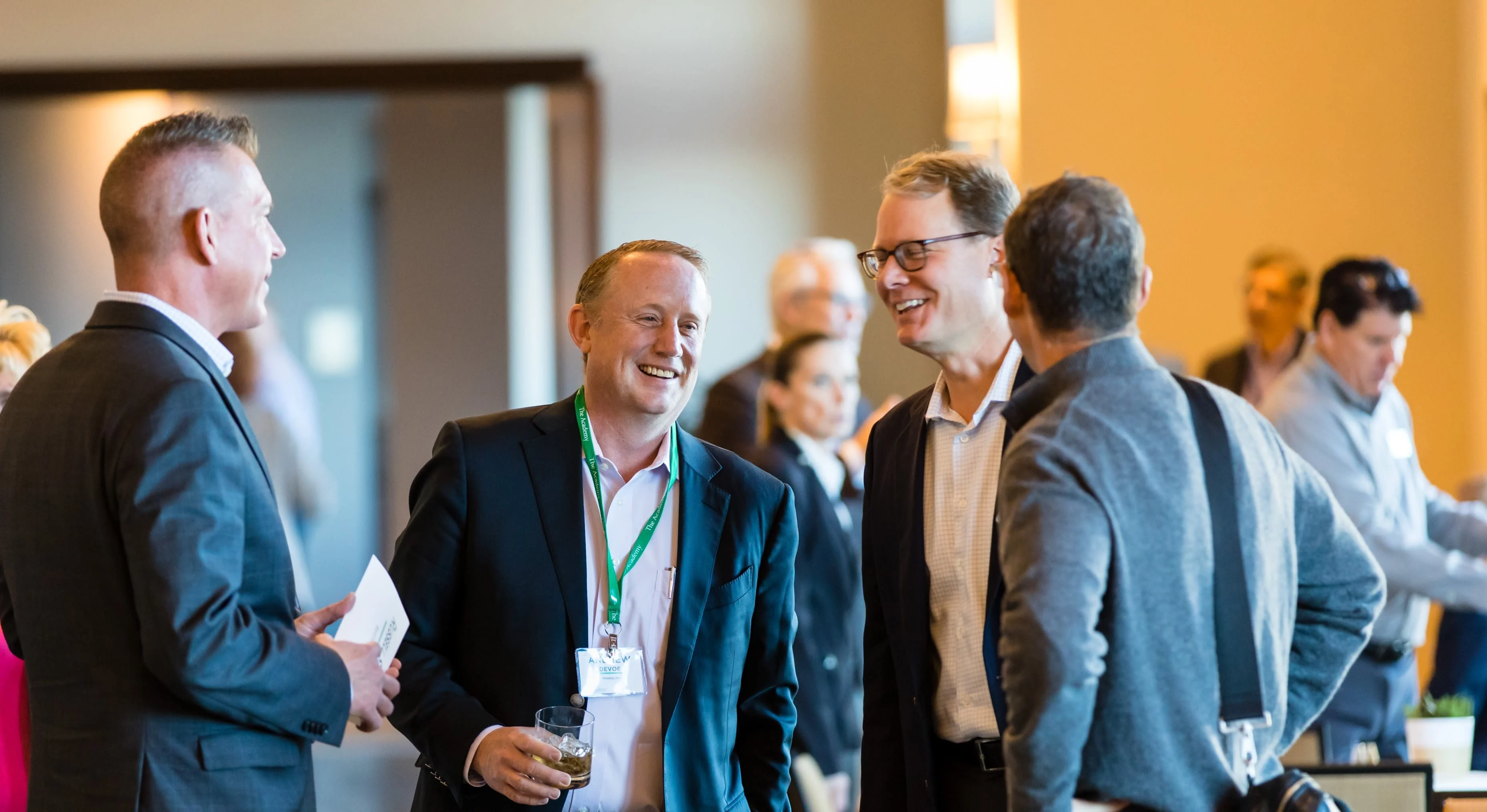 Group of professionally dressed individuals engaged in conversation at a networking event. Participants appear smiling and relaxed in a well-lit venue with others interacting in the background.







