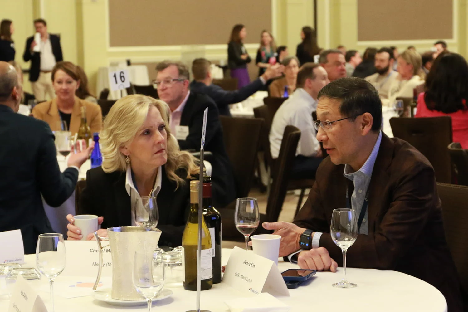 Two healthcare executives in business-casual attire sit at a banquet table in an event hall, conversing over drinks.