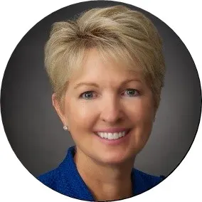 A professional circular headshot of a smiling woman with short blonde hair wearing a textured blue jacket, posed against a dark grey studio background.