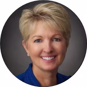 A professional circular headshot of a smiling woman with short blonde hair wearing a textured blue jacket, posed against a dark grey studio background.
