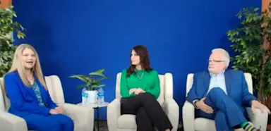 Three healthcare leaders seated in chairs against a blue backdrop during a discussion panel, featuring a woman in a blue suit speaking, another woman in green listening, and a man in a blue suit observing.