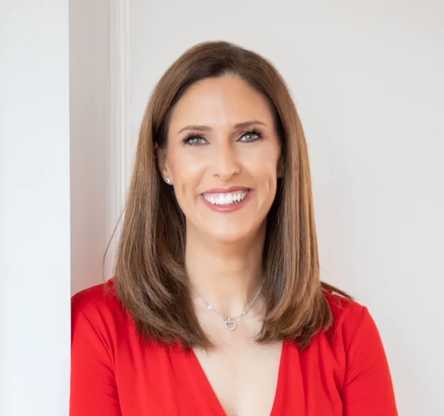 A woman with straight brown hair and light makeup smiles while standing against a white wall. She is wearing a red top and a silver necklace. The background is simple and softly lit.