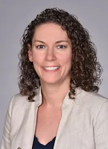 Professional headshot of a woman with curly brown hair, smiling and wearing a light beige blazer over a dark top, set against a plain gray background.