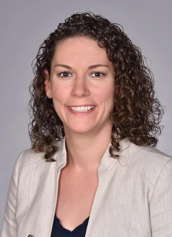 Professional headshot of a woman with curly brown hair, smiling and wearing a light beige blazer over a dark top, set against a plain gray background.