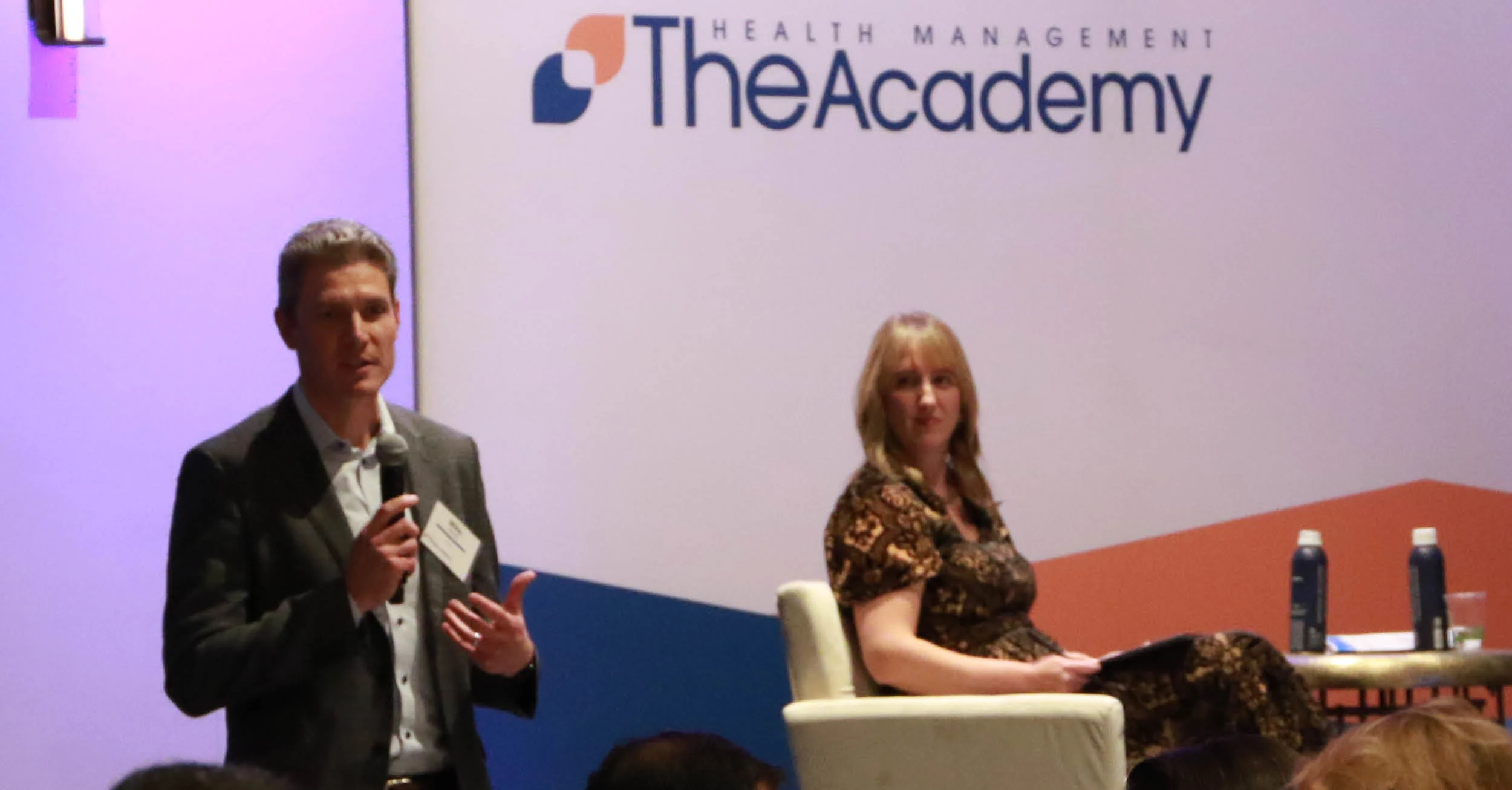 A panel presentation stage at a Health Management Academy event. On the left, a man in a grey suit jacket and white shirt stands while speaking into a microphone, gesturing with his hand. On the right, a woman sits in an armchair listening, dressed in a dark patterned dress. The background wall features a large logo that reads "The HEALTH MANAGEMENT Academy" above a modern geometric design in blue and orange.