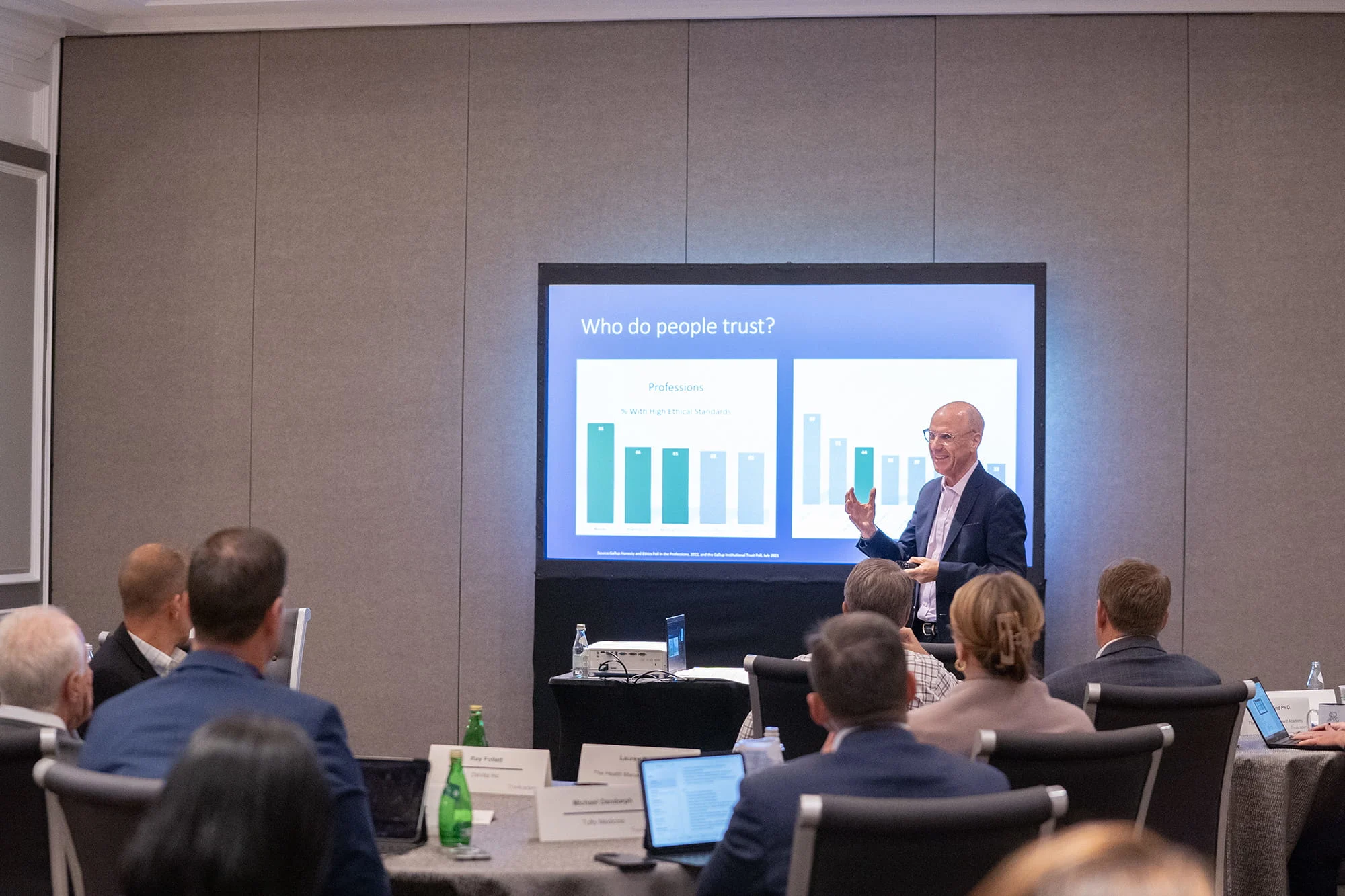 Presenter standing in front of a slide titled 'Who do people trust?' with bar charts, speaking to an audience seated at round tables in a conference room