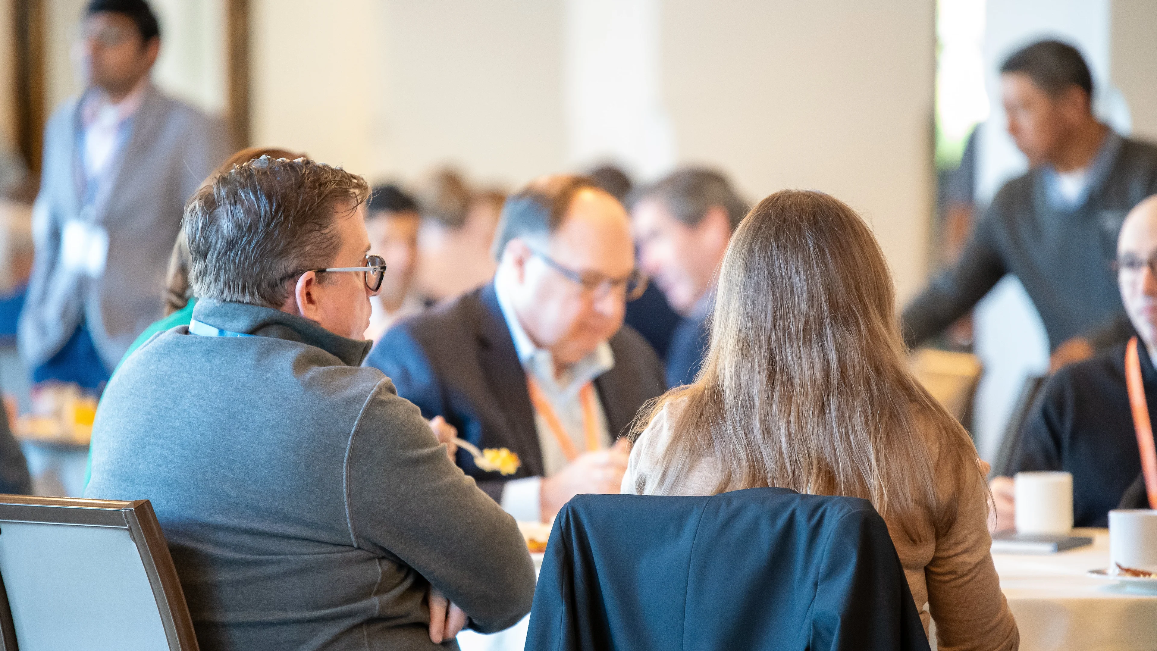 Attendees sit together at a round table during a conference meal, engaging in conversation, with others networking in the blurred background.