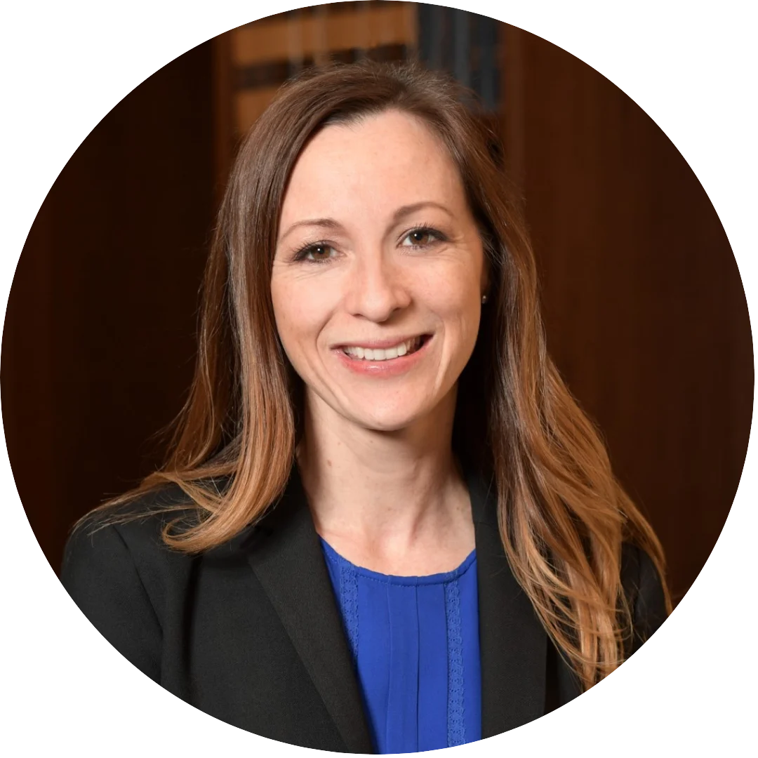 Smiling woman with long light brown hair wearing a black blazer and royal blue blouse, posed indoors with a warmly lit background and bookshelves behind her.
