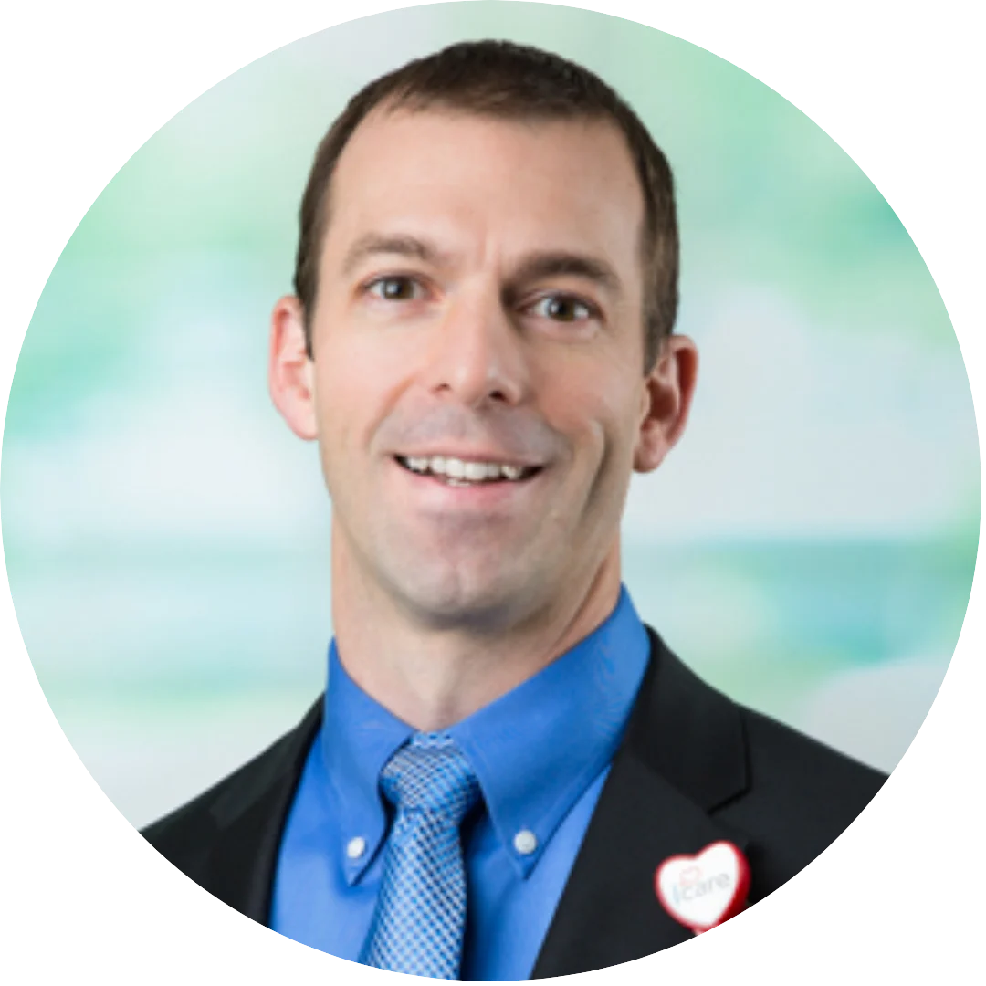 Smiling man wearing a black blazer, blue shirt, and patterned blue tie, with a heart-shaped badge on his lapel, posed against a soft, abstract green and blue background.