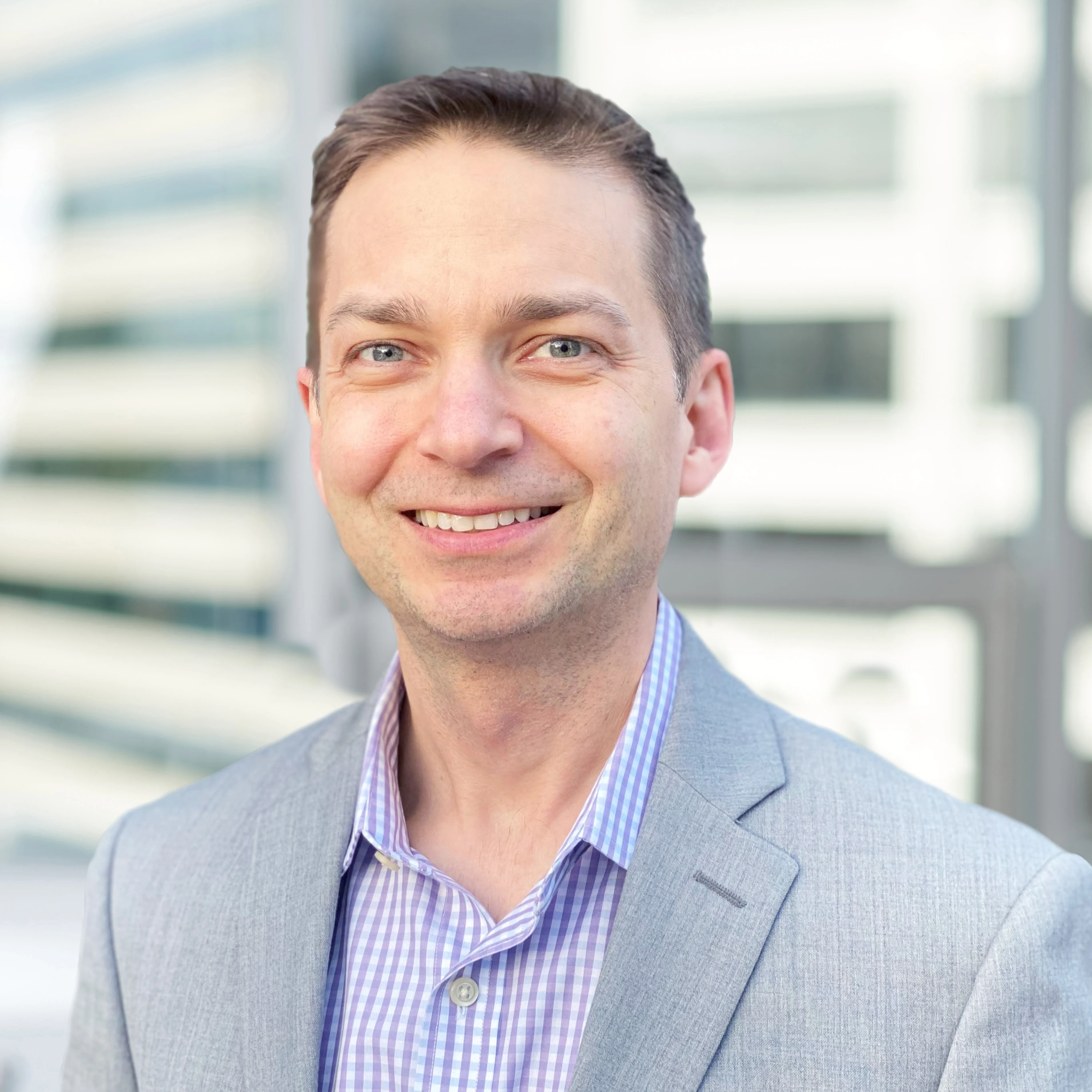 A professional headshot of a smiling man wearing a light gray blazer and a purple checkered shirt, standing in front of a blurred office building background with horizontal lines.