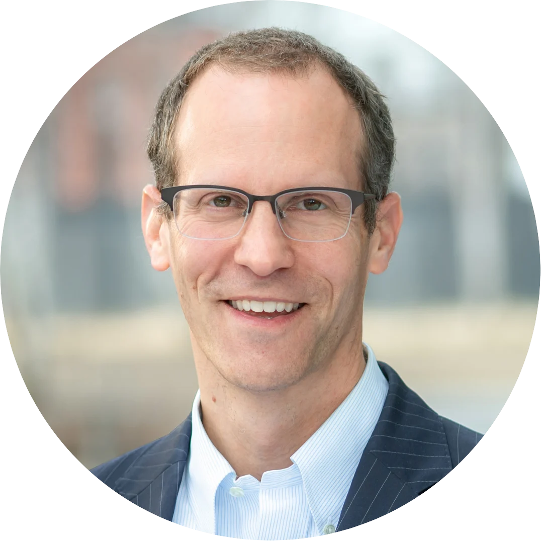 Smiling man wearing glasses, a light blue collared shirt, and a dark pinstripe blazer, photographed outdoors with a softly blurred cityscape in the background.