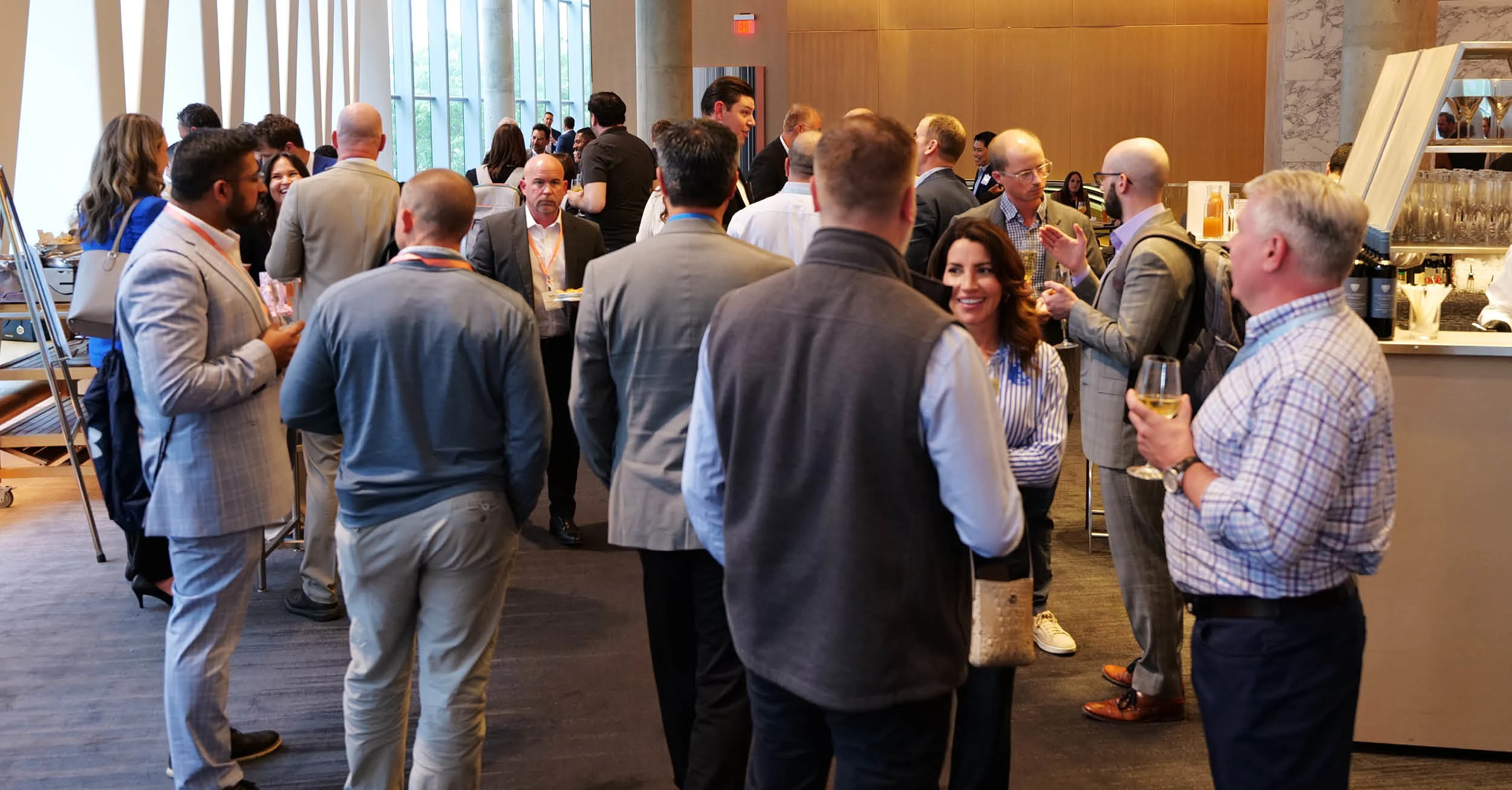 A crowded indoor networking reception with professionals mingling and talking in groups. Attendees are dressed in business casual attire, with some holding wine glasses and small plates of food. The venue features a modern design with large windows letting in natural light on the left, a tall wooden wall panel in the background, and a bar area set up on the right side.