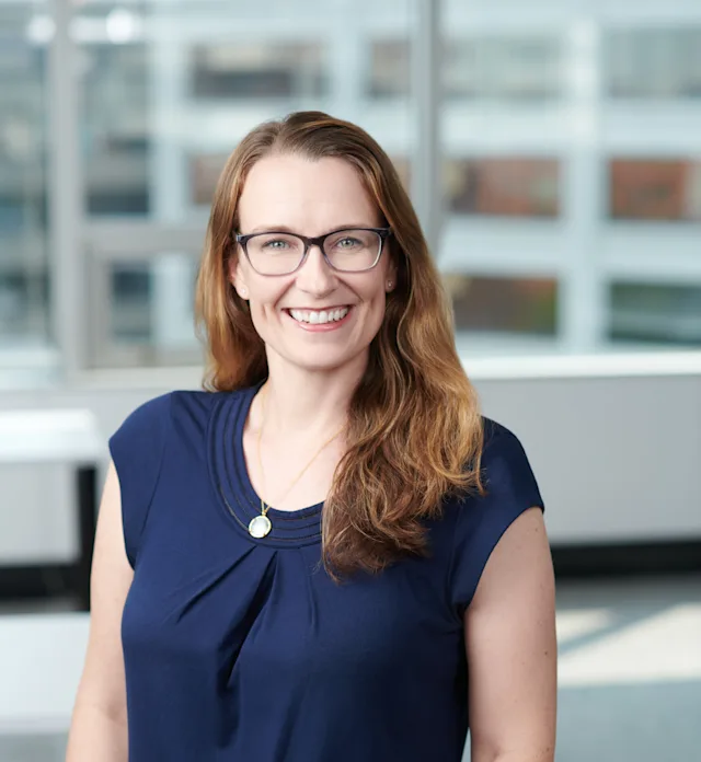 A woman with long wavy light brown hair and glasses smiles while wearing a navy-blue top with pleated detail and a pendant necklace. She stands in a brightly lit office with large windows in the background.