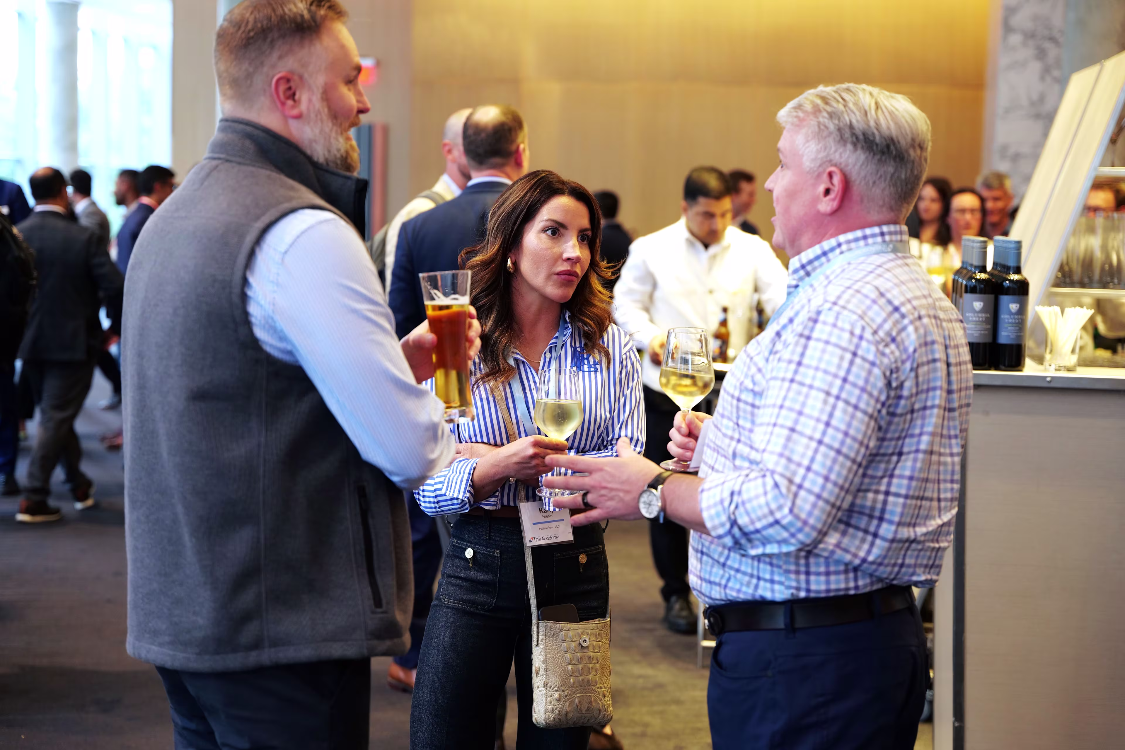 Three event attendees are seen engaged in conversation while holding drinks at a networking reception. The group includes two men and one woman standing near a bar area. The background shows a lively room filled with other professionals mingling in a modern, well-lit space.

