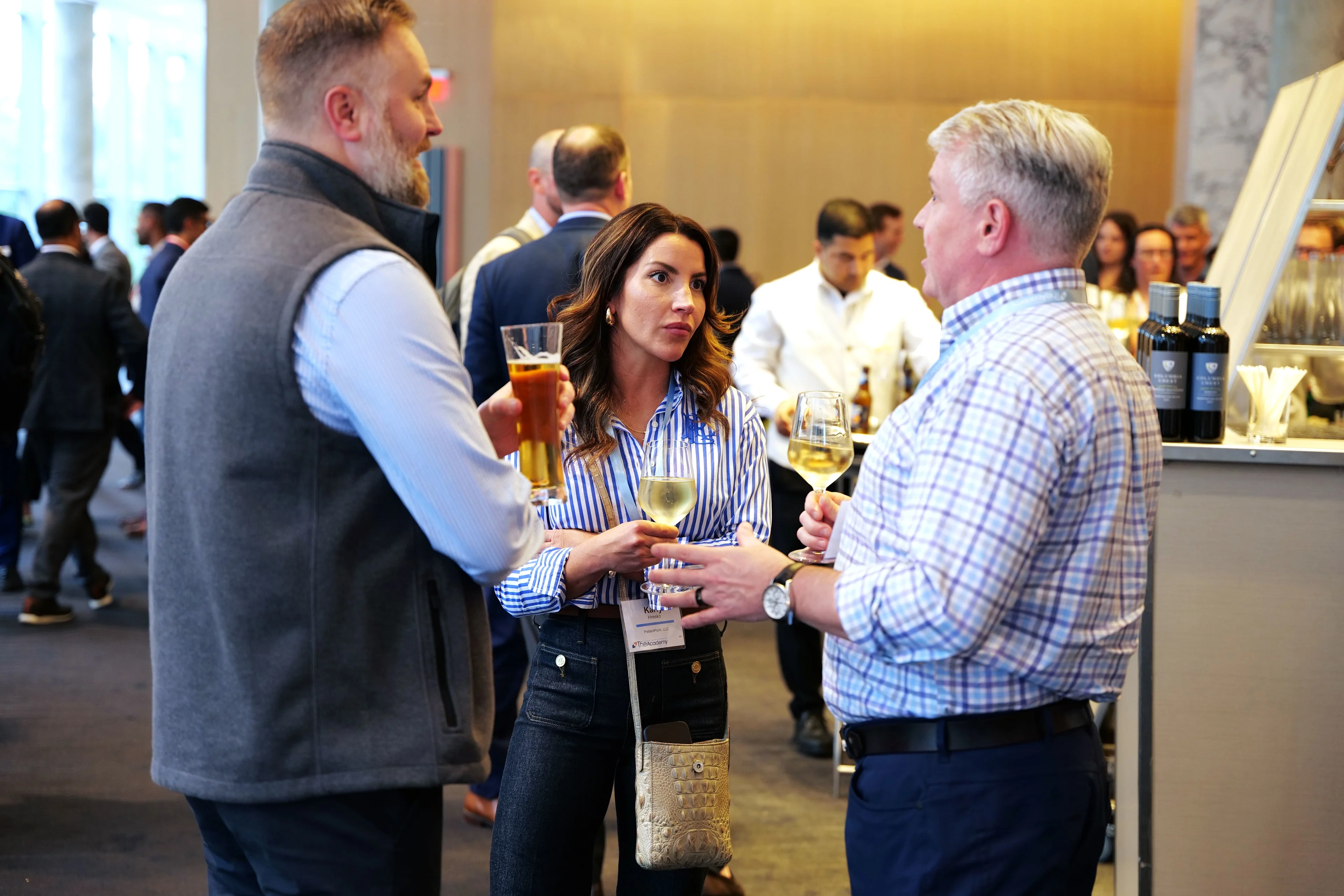 Three event attendees are seen engaged in conversation while holding drinks at a networking reception. The group includes two men and one woman standing near a bar area. The background shows a lively room filled with other professionals mingling in a modern, well-lit space.

