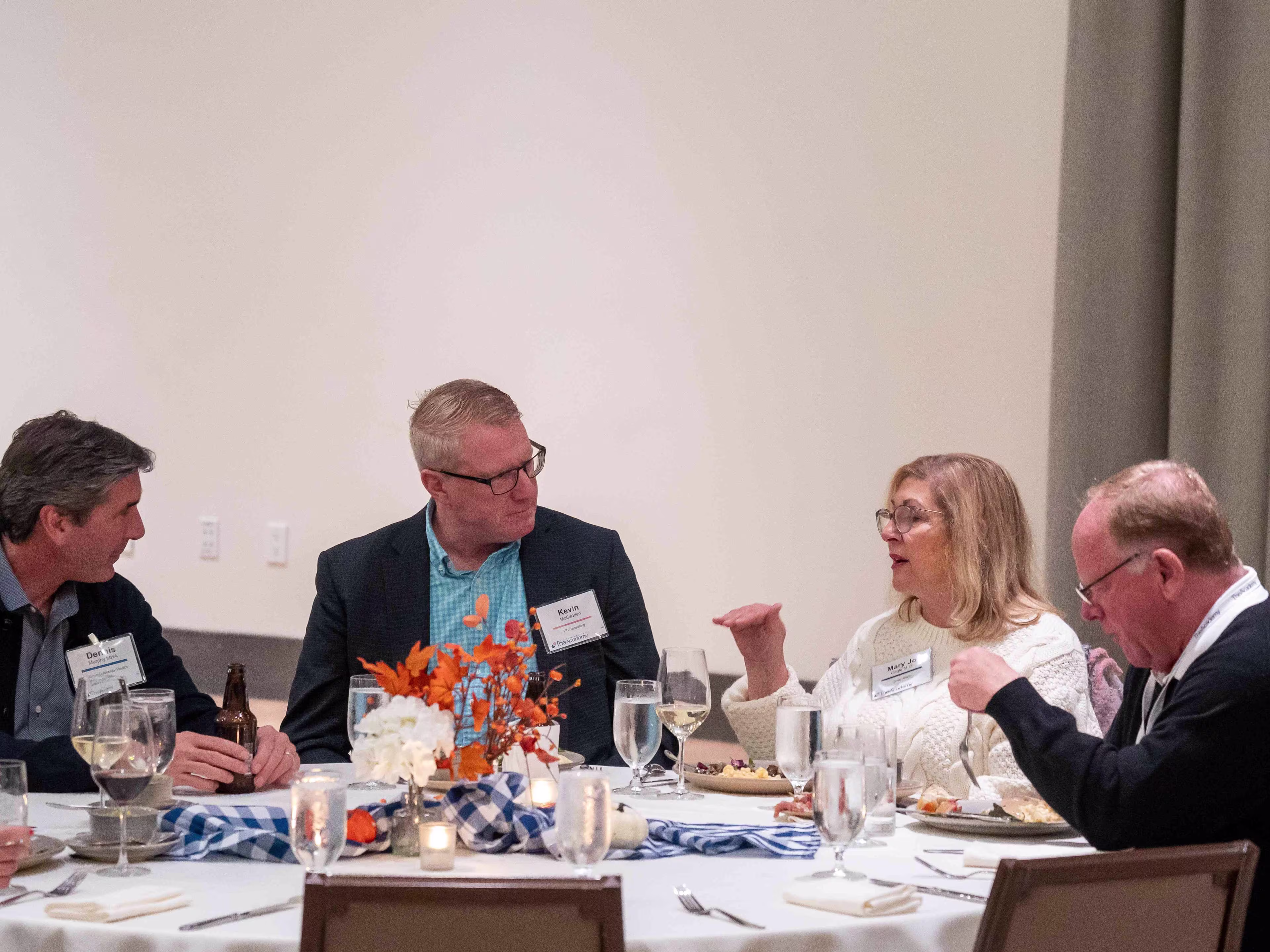 Executives seated at a round table engage in discussion during healthcare operations and administration conferences, with dinner settings and floral décor in the background.
