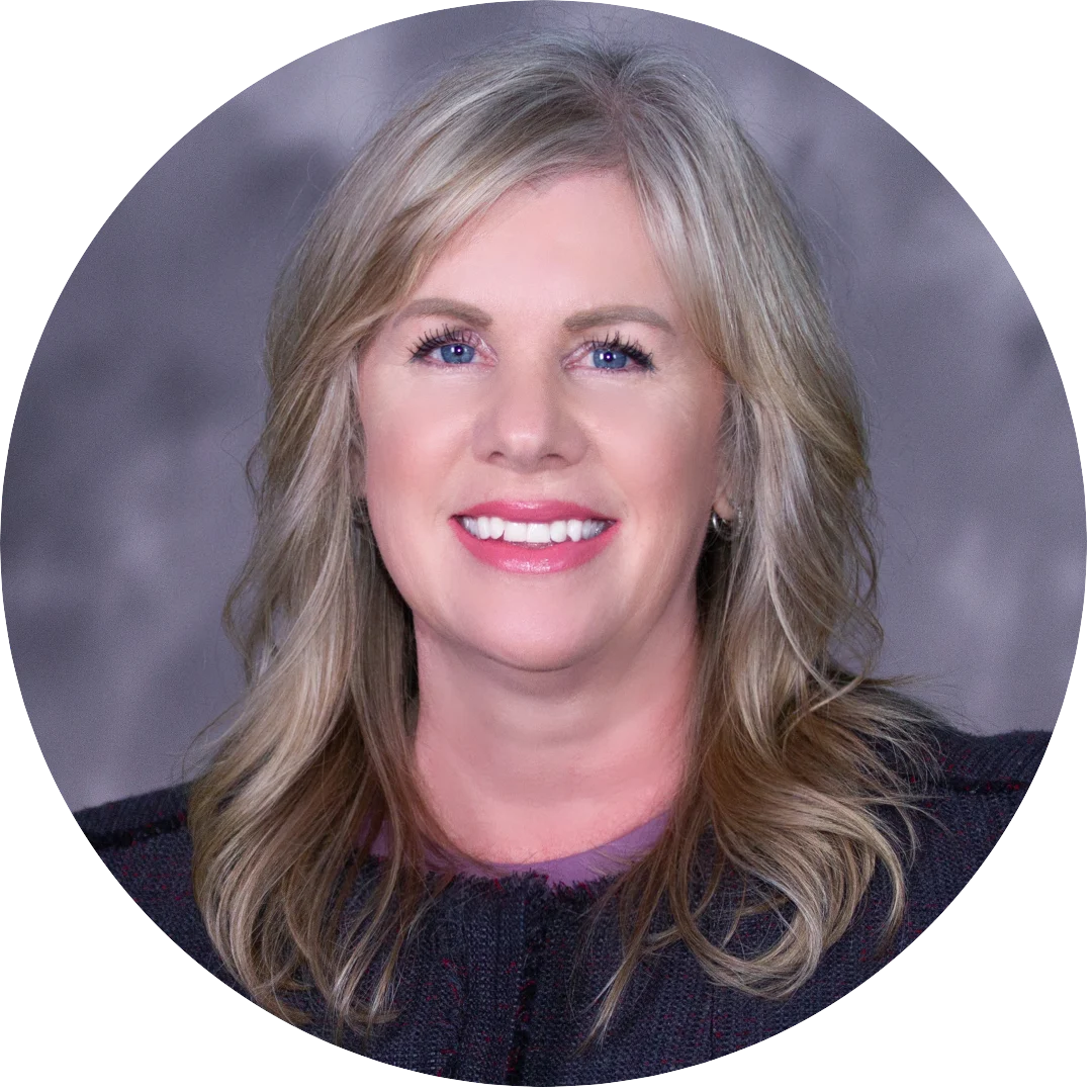 Smiling woman with long wavy blonde hair wearing a dark textured blazer, posed against a softly blurred purple-gray background.