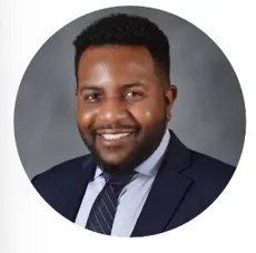 A man with short curly hair and a beard smiles while wearing a navy suit, white shirt, and striped tie, shown in a circular-cropped headshot against a gray background.