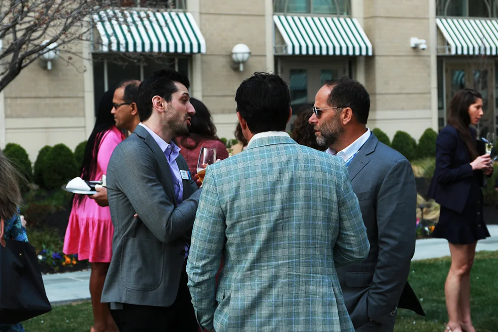 Group of professionally dressed individuals engaged in conversation at an outdoor networking event. People are holding drinks and plates while talking in small groups on a lawn in front of a building with striped awnings.
