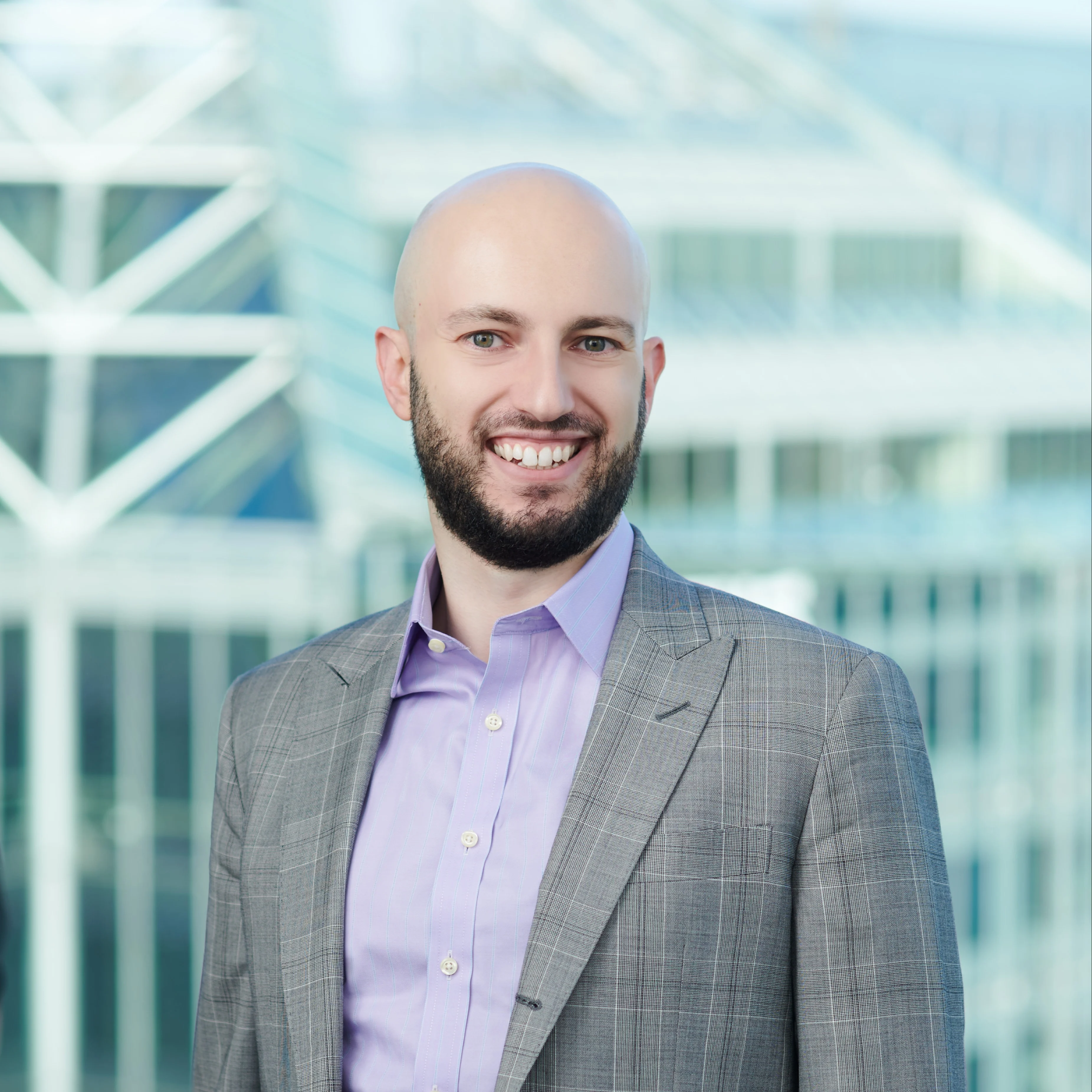A man in a light gray plaid suit and lavender shirt stands smiling in front of a modern glass office building, with geometric architectural elements in the background.