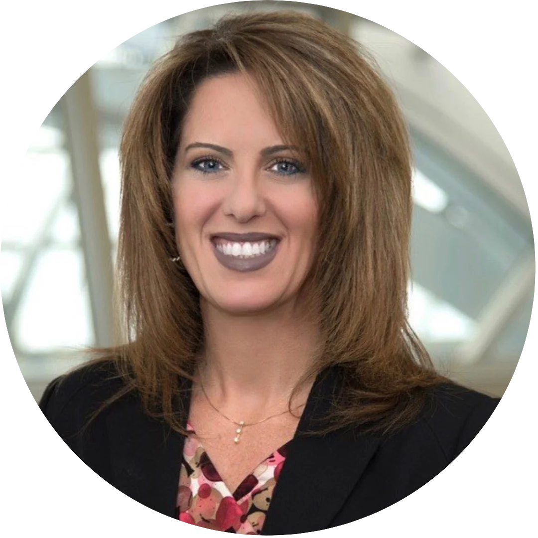 Woman with medium length layered brown hair wearing a black blazer over a floral blouse, smiling in a well-lit indoor environment with a modern architectural background.
