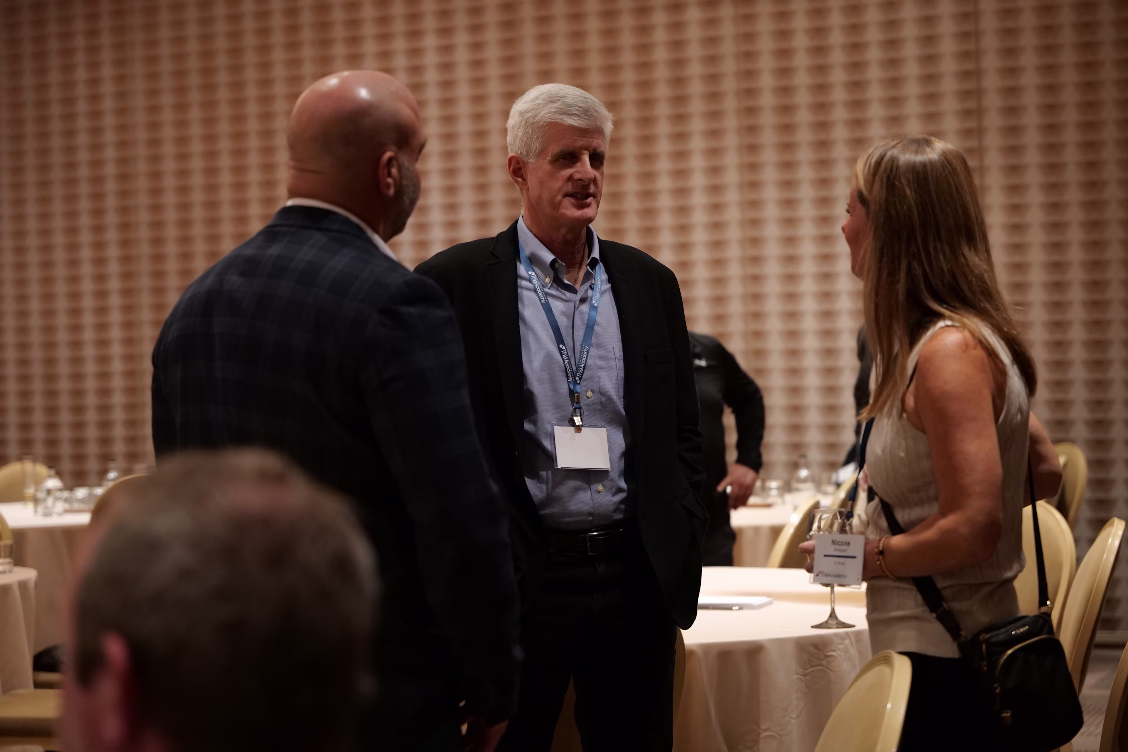 Three attendees stand and converse in a dimly lit conference room with round tables and neutral decor. A man in a dark blazer and a woman with a lanyard engage in conversation with another attendee, suggesting a casual networking moment during a break in the event.

