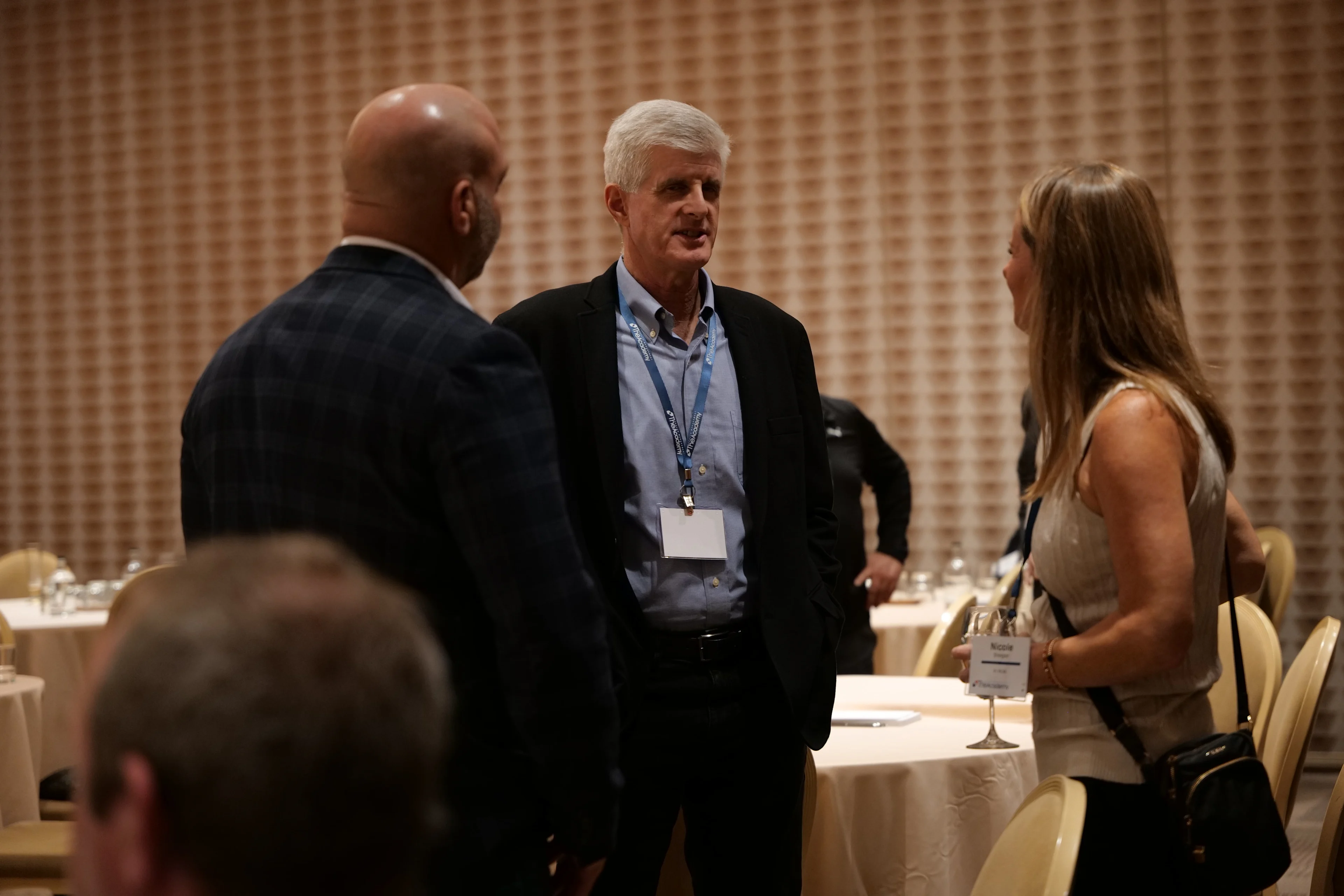 Three attendees stand and converse in a dimly lit conference room with round tables and neutral decor. A man in a dark blazer and a woman with a lanyard engage in conversation with another attendee, suggesting a casual networking moment during a break in the event.

