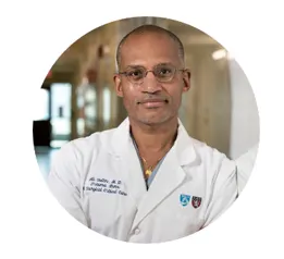 A man wearing glasses and a white medical coat with hospital logos stands confidently in a hallway, arms crossed and facing the camera.