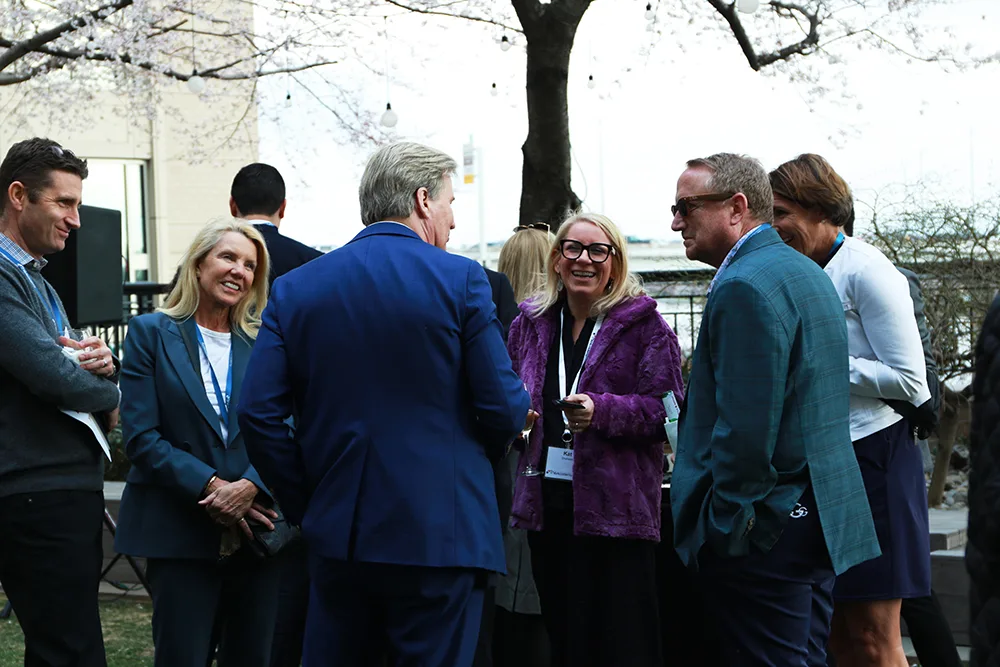 Group of professionals engaged in conversation at an outdoor gathering. Most are wearing business attire, and the setting features a blooming tree and a building in the background. Attendees appear to be enjoying light conversation in a relaxed, social atmosphere.