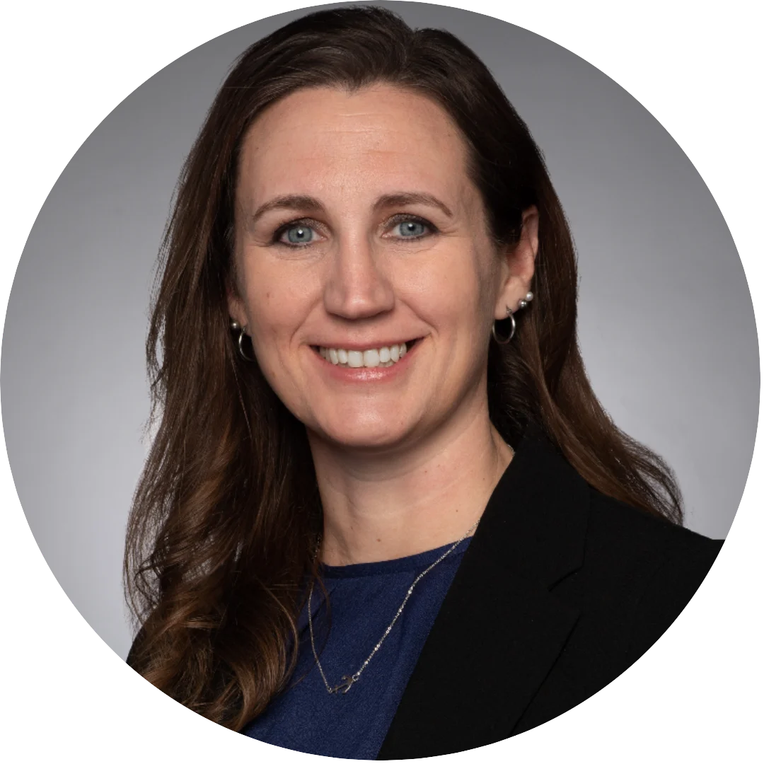 Smiling woman with long brown hair wearing a navy blouse, black blazer, and delicate necklace, posed against a neutral gray background.