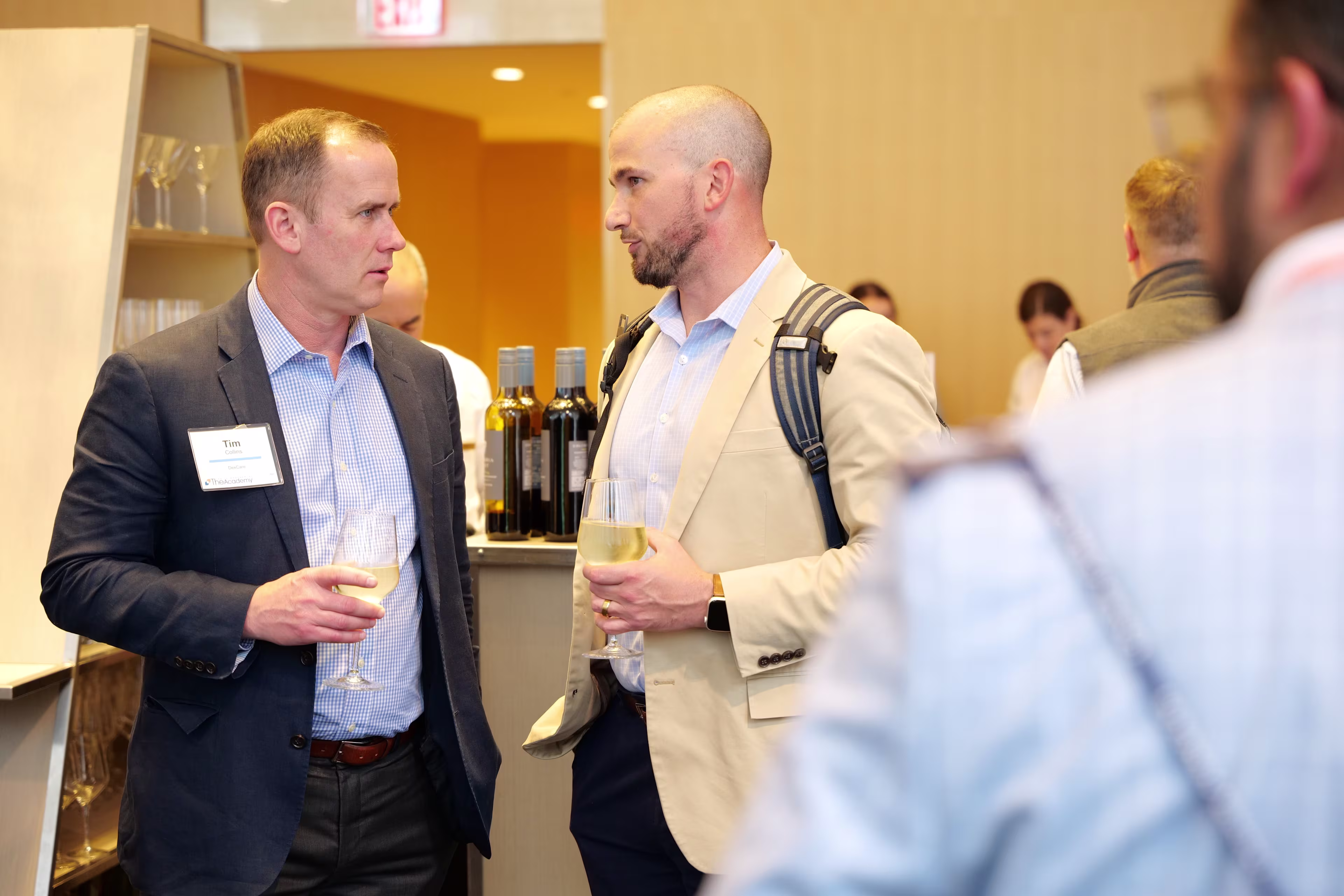 Two men converse while holding drinks during a networking event, with bottles and other attendees visible in the background.

