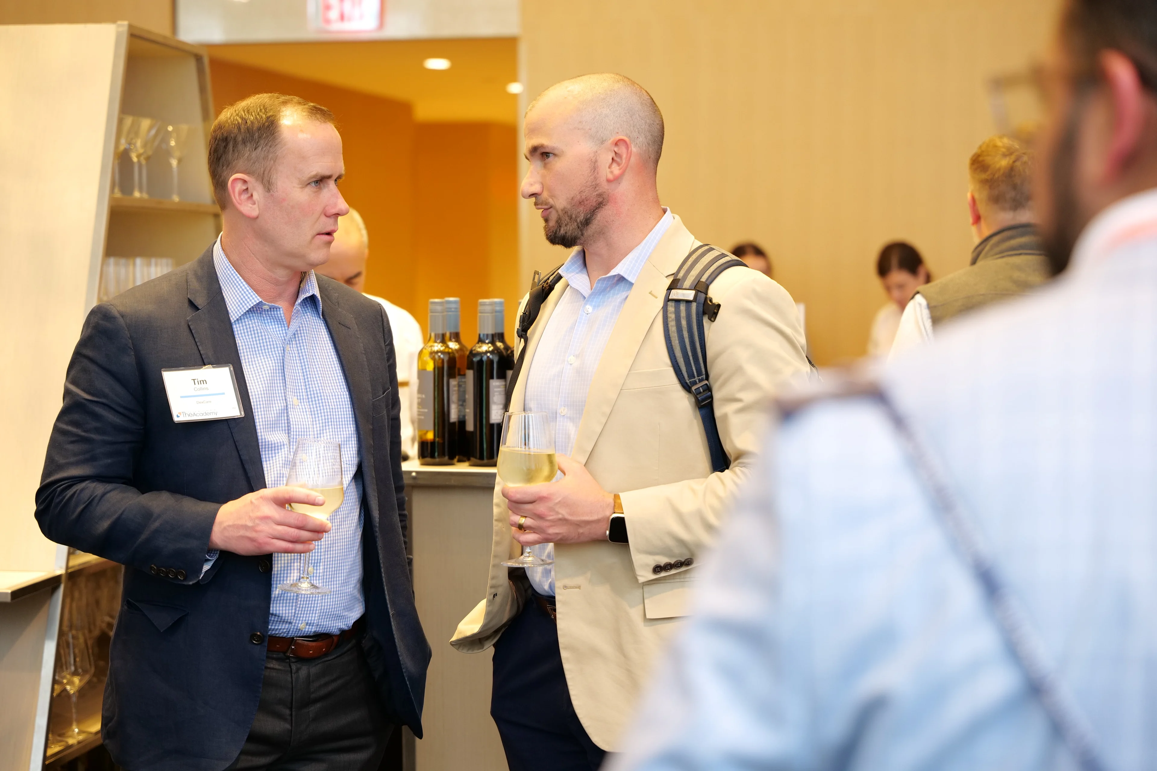 Two men converse while holding drinks during a networking event, with bottles and other attendees visible in the background.
